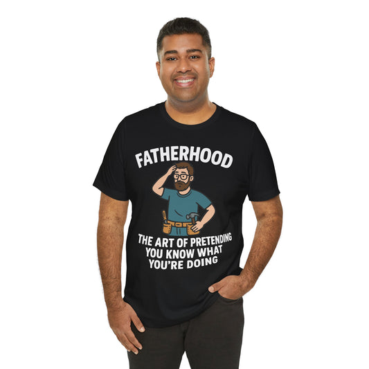 Black t-shirt featuring a cartoon dad scratching his head with tools on his belt and the quote "Fatherhood: The Art of Pretending You Know What You're Doing."
