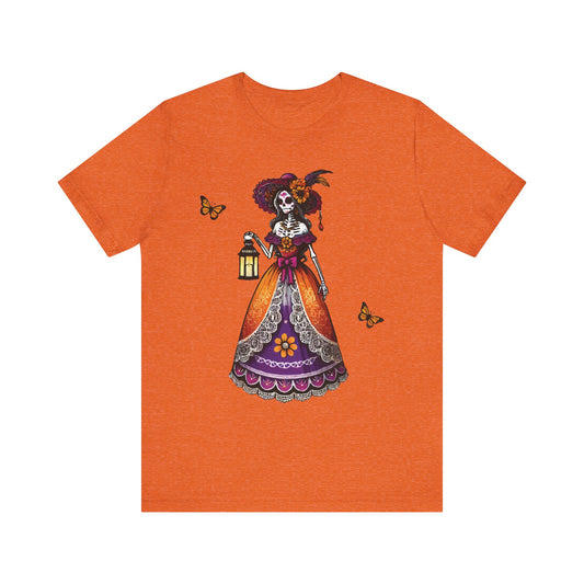 Heather orange t-shirt with a bold Día de los Muertos design of La Catrina skeleton lady in a vibrant dress holding a lantern, with monarch butterflies.


